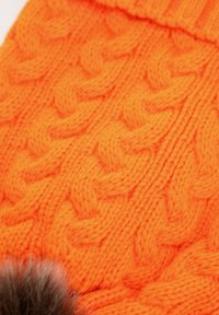 Cappello arancione lavorato a maglia con un motivo a cavo spesso e testurizzato e un pon pon in pelliccia marrone decorativo sul bordo inferiore.
