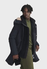 Manteau rembourré bleu marine avec un col en polaire noir et beige, fermeture éclair à l'avant et poches latérales. Le modèle porte une chemise verte en dessous.