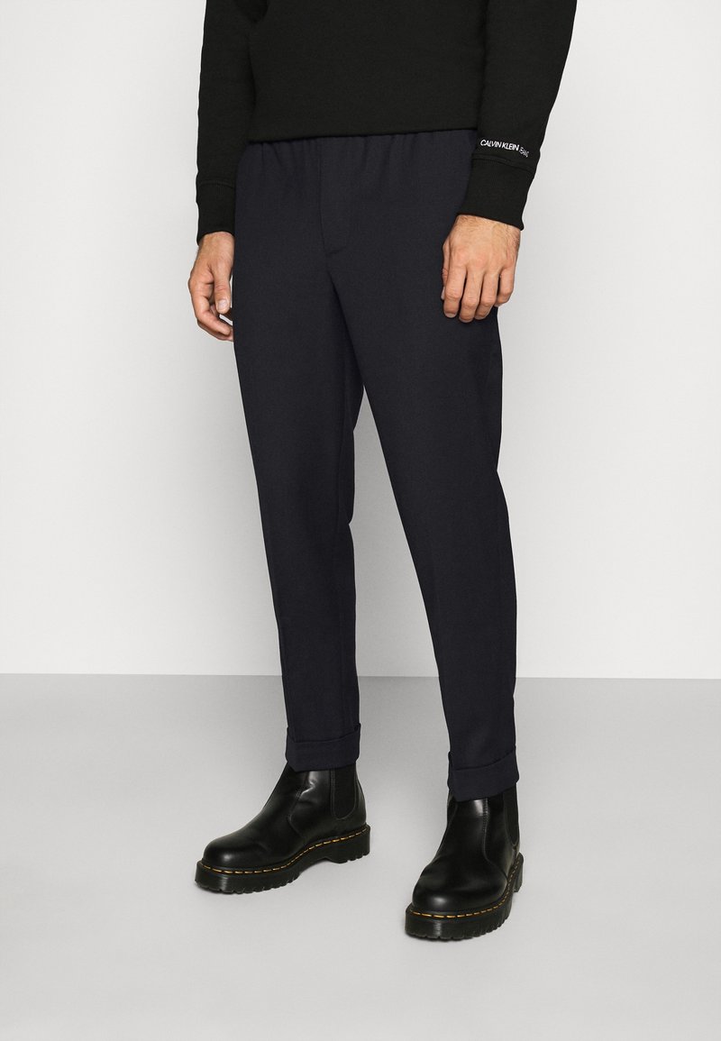 Homme portant une chemise noire à manches longues, un pantalon bleu marine avec revers, et des bottines en cuir noir, debout sur un sol gris contre un mur blanc.