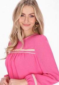 Maglietta rosa con maniche lunghe, caratterizzata da un tessuto testurizzato e strisce decorative color crema. Il design include sezioni arricciate e polsini elastici.
