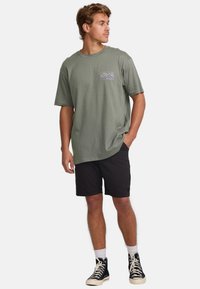 Olivgrünes T-Shirt mit kurzen Ärmeln, ausgestattet mit einem grafischen Design auf der Vorderseite. Kombiniert mit schwarzen Shorts und schwarzen High-Top-Sneakern.