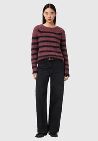 Femme aux cheveux noirs lisses portant un pull pelucheux violet et noir à rayures, un jean noir à jambes larges et des chaussures noires, sur un fond gris.