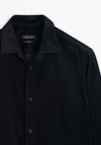 Sort kordfløjlsskjorte med spidse krave, lodret ribbet tekstur og fem knapper foran. Etiketten siger "Massimo Dutti."