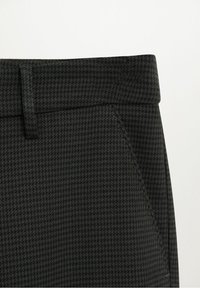 Gros plan d’un pantalon gris foncé à motif pied-de-poule avec ceinture, passants et couture de poche latérale sur un fond clair.
