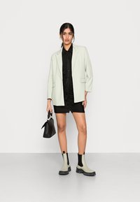Blazer oversized verde claro sobre um vestido preto drapeado, combinado com botas de tornozelo beje e pretas e uma pequena mala preta.