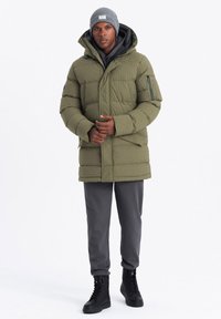 Parka aislante de color verde oliva con textura acolchada, cremallera frontal, bolsillos laterales y capucha. Complementada con un gorro gris y pantalones oscuros.