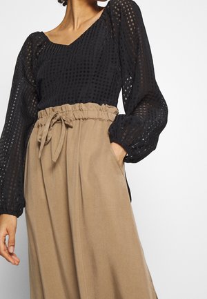 Vrouw draagt een zwarte doorschijnende blouse met lange mouwen met geometrisch patroon en beige broek met hoge taille, strikceintuur en elastische tailleband.