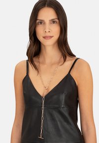 Lang, sammenhængende kædehalssmykke i rosaguld, med et minimalistisk design og en T-bar detalje. Båret over en sort lædertop.