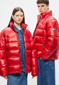 Κόκκινα μπουφάν puffer με γυαλιστερό φινίρισμα, με ψηλό γιακά, φερμουάρ μπροστά και πλαϊνές τσέπες. Φοριούνται πάνω από πουκάμισα τζιν με σχέδια.