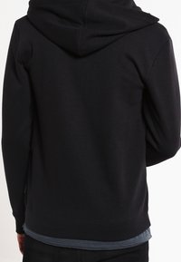 Felpa nera con cappuccio regolabile, polsini e orlo a costine, caratterizzata da una texture liscia e un design minimalista. Strati visibili con una maglietta grigia.