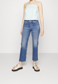 Svetle modrý zkrácený top bez rukávů s texturovaným vzorem, spárovaný s modrými džíny s vysokým pasem a bílými sandály s černými podrážkami.