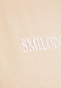 Beżowy materiał o gładkiej teksturze z wytłoczonym na środku słowem "SMILODO" na biało, wyraźnie widocznym.