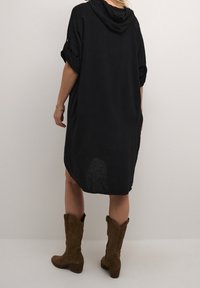 Svart linneklänning med huva, korta uppvikta ärmar och sidofickor, matchad med bruna mocka knähöga stövlar. Strukturerad tyg synlig.