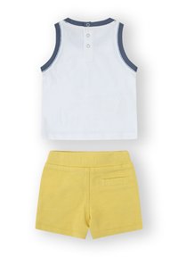 Top sin mangas blanco con ribete azul oscuro y botones en la parte trasera, combinado con pantalones cortos de algodón amarillo que tienen un bolsillo en la parte trasera.