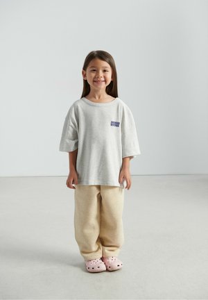 Siva oversized majica s logotipom "American Vintage", bež jopič hlače i ružičaste klompe s rupama za ventilaciju, stojeći na glatkom podu.