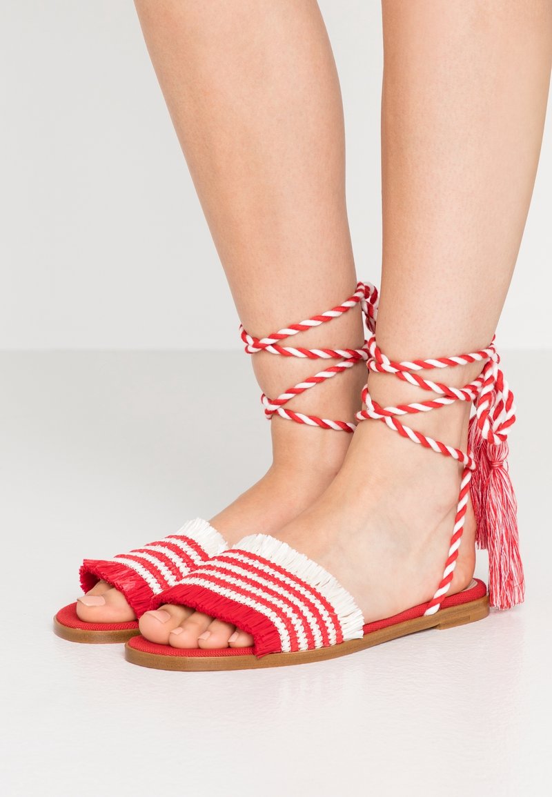 Pieds portant des sandales plates rayées rouge et blanc avec des brides à franges et des liens de cheville torsadés rouges et blancs avec des pampilles.