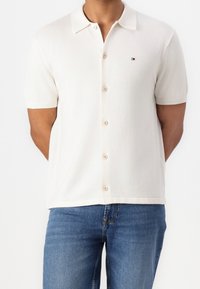 Koszulka polo z krótkim rękawem w kolorze kremowym, wykonana z miękkiego materiału, z zapięciem na guziki z przodu oraz małym logo na lewej piersi.