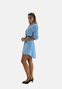 Light blue shirt dress with a collared neckline and rolled sleeves, cinched at the waist with a black belt, paired with black slingback heels.