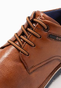 Chaussure en cuir marron avec une finition texturée, dotée de lacets crème, d'un bout lisse et d'une semelle en caoutchouc sombre. Mise en valeur par une étiquette noire.