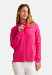 Haut de sweatshirt rose zippé en tissu doux, avec un col haut, des poches avant et des accessoires en or. Texte "FREDDY" imprimé sur la poitrine.