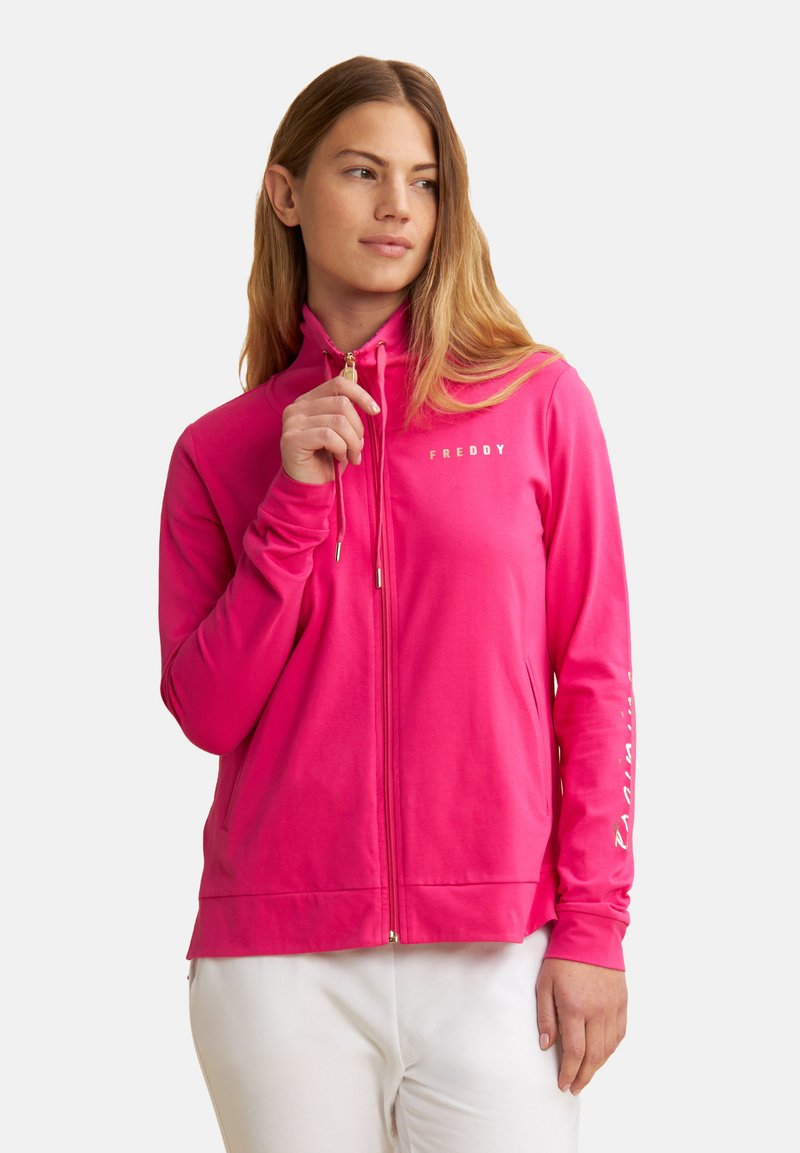 Haut de sweatshirt rose zippé en tissu doux, avec un col haut, des poches avant et des accessoires en or. Texte "FREDDY" imprimé sur la poitrine.
