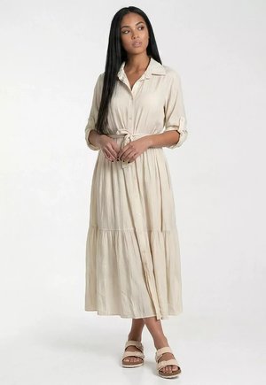Kobieta z długimi ciemnymi włosami ubrana w beżową midi sukienkę na guziki z długimi rękawami, wiązaną w talii, oraz beżowe sandały, stojąca na białym tle.