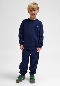Mörkblå sweatshirt och joggers, i mjukt tyg, med en liten logotyp på bröstet och elastiska muddare, tillsammans med gröna sneakers.