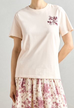 Hellrosa Baumwoll-T-Shirt mit kurzen Ärmeln und floralem Druck in dunkelrot auf der linken Seite. Weiche Textur, entspannter Schnitt.