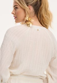 Top blanco texturizado con un patrón de cuadrícula, que presenta un detalle de botón dorado en la parte posterior y adornado con pendientes florales dorados.