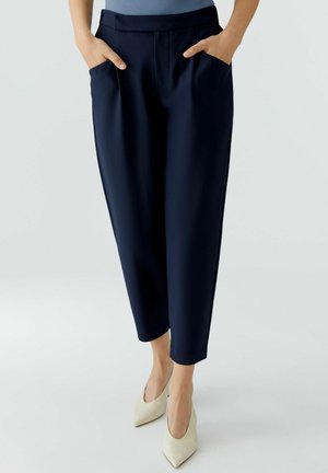 Femme portant un pantalon marine fuselé aux chevilles, mains dans les poches, et des talons beige à bout pointu sur fond clair.