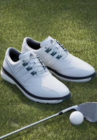 Bílé kožené golfové boty s hladkým povrchem, třemi námořnickými pruhy a texturovanou podrážkou. Kousek dál se nachází golfová hůl a míček na zelené trávě.