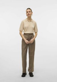 Polo beige con maniche corte, tre bottoni, abbinato a pantaloni marroni a vita alta e scarpe nere stringate. Tessuto di texture liscia.