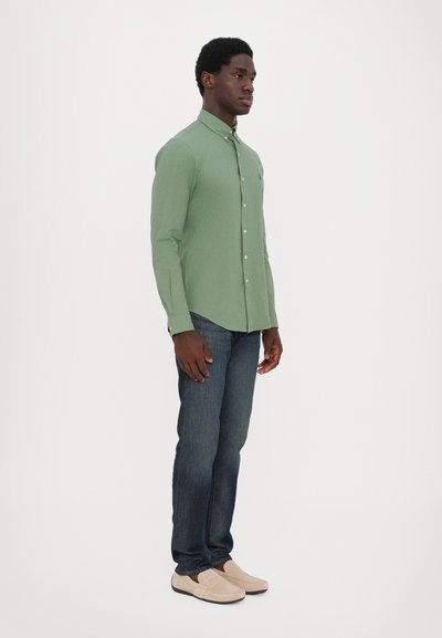 Chemise verte à manches longues avec un col boutonné, portée avec un jean foncé et des chaussures slip-on beige clair, mettant en avant un look décontracté.
