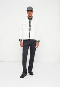 Biała dżinsowa kurtka na zielonej koszuli w kratę, połączona z szarymi spodniami i czarnymi loafersami. Model nosi szary kapelusz. Prosty, zorganizowany design.