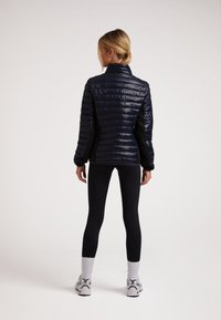 Marineblaue Steppjacke mit vertikaler Steppung, hohem Kragen, figurbetontem Design, kombiniert mit schwarzen Leggings und weißen Sportschuhen.