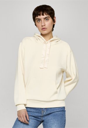 Giovane con capelli corti e scuri che indossa una felpa crema con cappuccio e jeans blu, in piedi davanti a uno sfondo grigio uniforme.