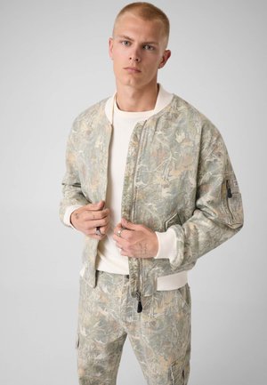 Jeune homme aux cheveux blonds courts portant une veste camouflage de couleur claire et un pantalon assorti sur une chemise blanche, debout devant un fond gris uni.