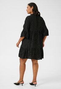 Vestido negro con rayas metálicas verticales, corsé ajustado, falda acampanada de capas y mangas de campana, combinado con mules negros de tacón.