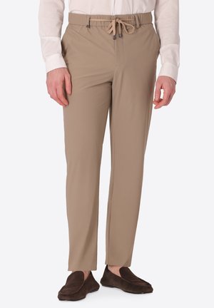 Uomo che indossa pantaloni beige con coulisse abbinati a una camicia bianca a maniche lunghe e scarpe slip-on marroni scure, in piedi dritto.