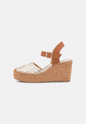 Wedge heel sandal with braided jute sole, metallic silver toe cap, and brown adjustable strap. Features textured fabric and rounded shape.