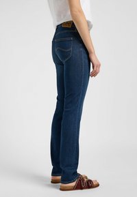 Jeans de mezclilla azul oscuro con una altura media, corte ajustado, que cuentan con costuras naranjas y una etiqueta de cuero marrón en la cinturilla.