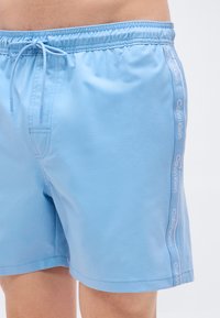 Ανοιχτό μπλε μαγιό βερμούδα με ελαστική μέση και κορδόνι. Διαθέτει λευκό λογότυπο στο πλάι και υφασμάτινη υφή, λεία στην αφή.