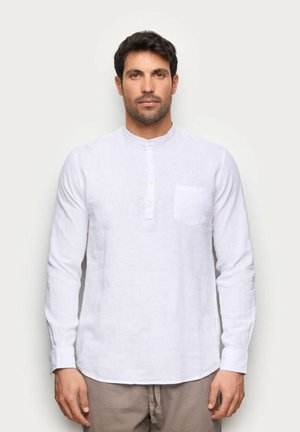 Mężczyzna o ciemnych włosach i brodzie, ubrany w białą, długorękawową koszulę z guzikami oraz jasnobrązowe spodnie, stojący na tle jednolitego jasnego tła.