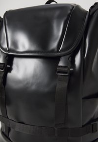 Zaino nero realizzato in materiale lucido con chiusura a pattina, spallacci regolabili e hardware con fibbia. Caratterizzato da una texture liscia e un design minimale.