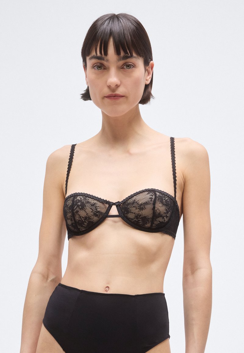 Melns mežģīņu balkonetveida krūšturis ar regulējamām viļņotām lencēm, ar polsterētām krūzītēm un centrālu loku uz vienkārša fona.