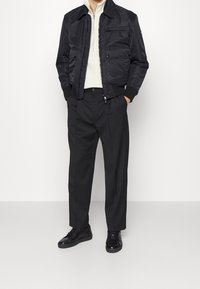 Svart bomberjacka över en krämfärgad tröja, i kombination med svarta byxor och svarta sneakers. Jackan har fickor och ribbade mudd.