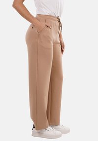 Pantaloni della tuta beige con vita elasticizzata e coulisse, tasche laterali e vestibilità comoda, abbinati a sneaker bianche, mostrati di lato.