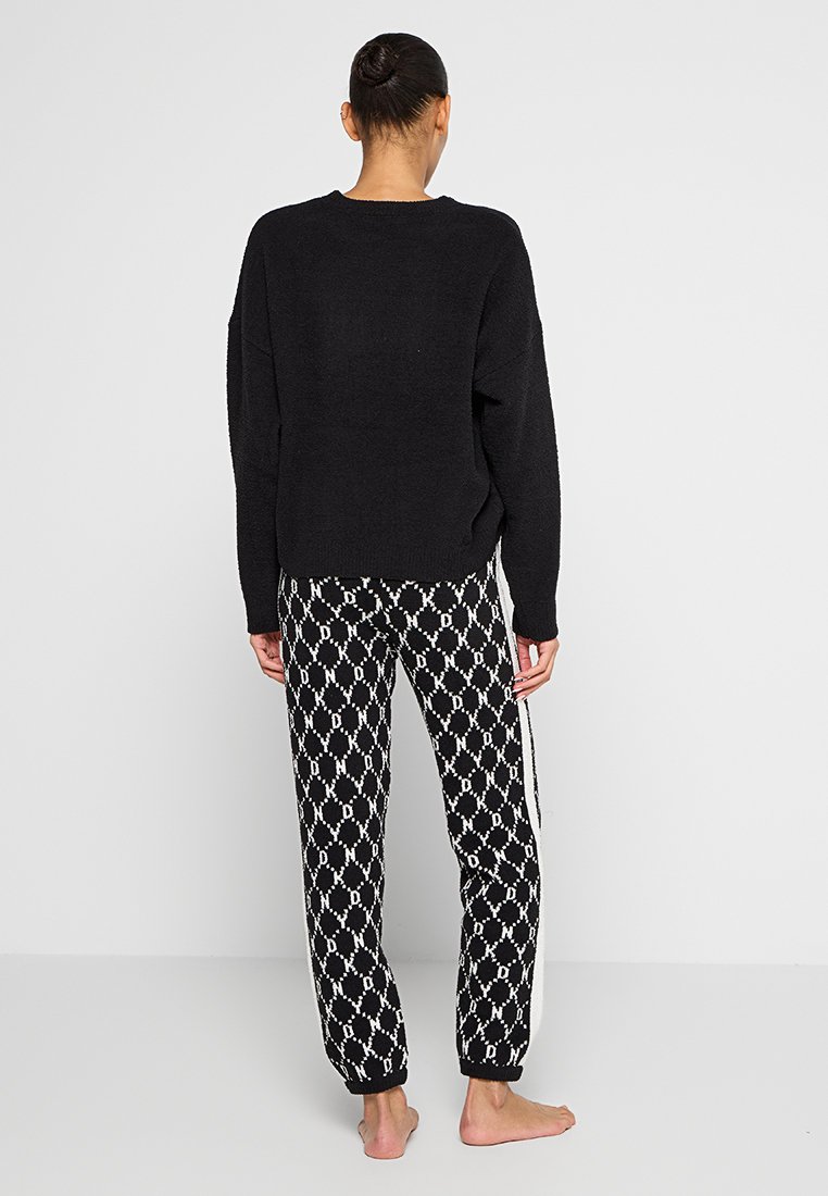 Schwarzer Strickpullover und gemusterte schwarz-weiße Hose mit einem sich wiederholenden Rautendesign und Seitenstreifen. Modell von hinten gezeigt.