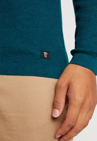 Närbild av en hand som vilar vid beige byxor, iklädd en strukturerad turkos tröja med en liten svart tygmärkning nära fållen.