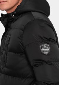 Giacca a vento nera con cappuccio, caratterizzata da una finitura testurizzata, tasca con zip e un'applique con logo sulla manica sinistra.